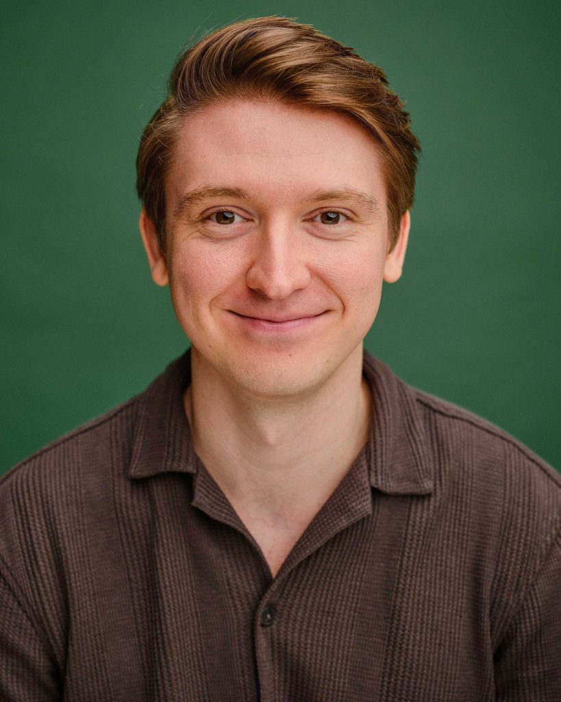 A young man with light skin and short brown hair is smiling at the camera, wearing a brown textured shirt, with a plain green background.